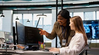 Eine freundliche leitende Ingenieurin betreut eine gut gelaunte Nachwuchsentwicklerin an einem Arbeitsplatz im Büro und überprüft die Kennzahlen des KI-Systems. Eine zufriedene afroamerikanische Managerin führt einen Praktikanten in die Codeüberprüfung mithilfe von maschinellem Lernen ein
