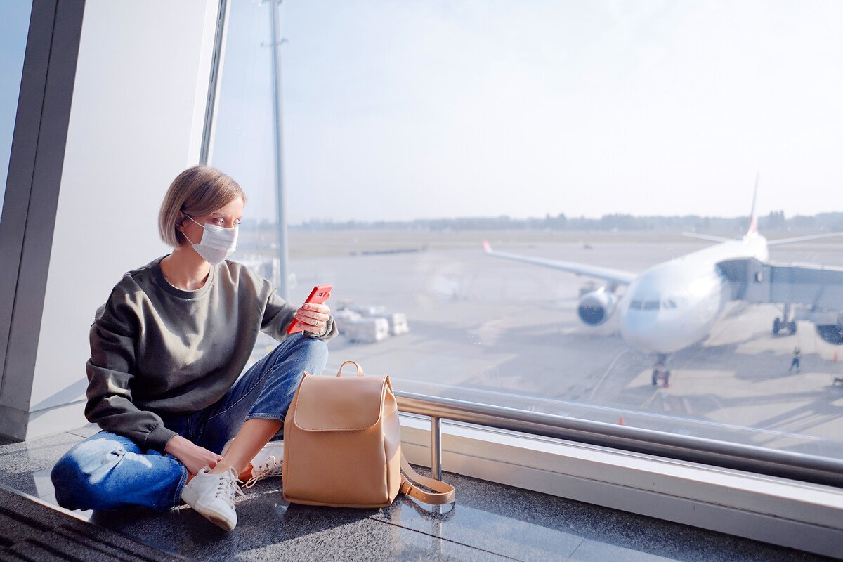 Eine junge Frau sitzt während der Corona-Schutzmaßnahmen mit einer Schutzmaske am Flughafen und sieht auf ihr Smartphone.