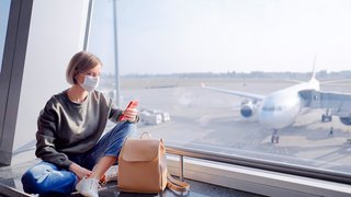 Eine junge Frau sitzt während der Corona-Schutzmaßnahmen mit einer Schutzmaske am Flughafen und sieht auf ihr Smartphone.