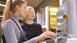 Eine junge Auszubildende erhält Unterricht von ihrer Ausbilderin. Beide Frauen arbeiten als Ingenieurinnen für computergesteuerte numerische Steuerungen. Sie sind an ihrem Hightech-Arbeitsplatz zu sehen.