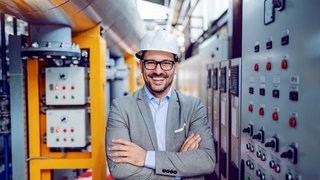 Smiling handsome caucasian businessman in gray suit and with helmet on head standing with arms folded next to dashboard. Power plant interior.